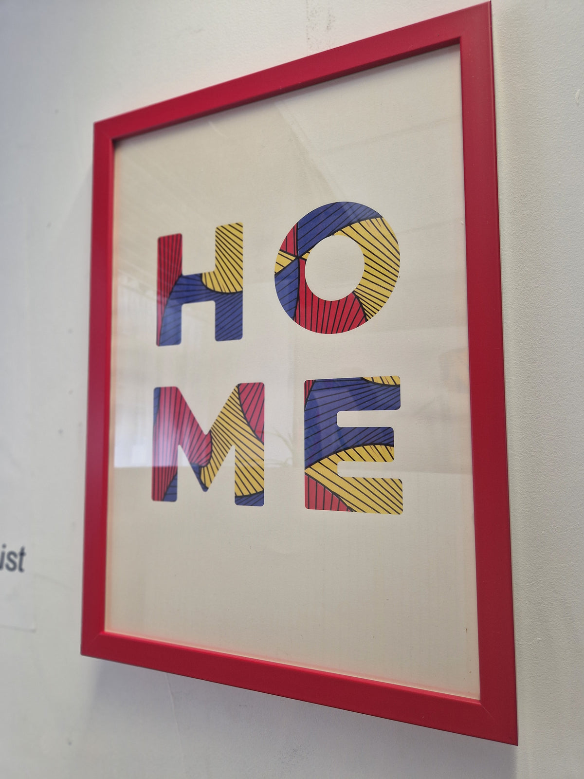 Framed wall art featuring the word "HOME" in large multicolored patterned letters inside a bright red frame, mounted on a white wall.