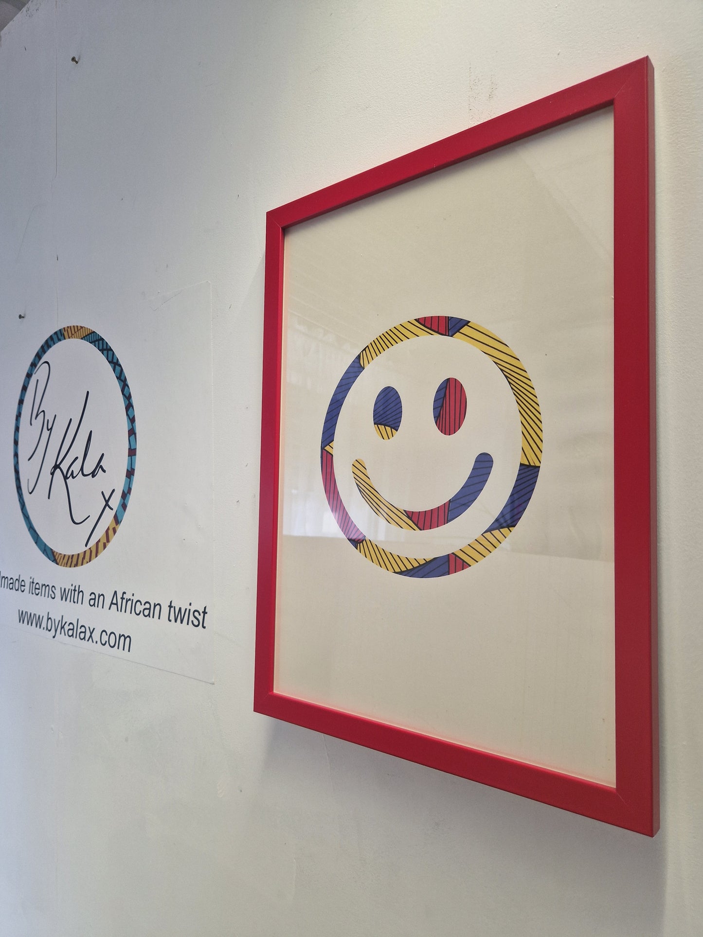 A framed art print featuring a smiley face made from colorful hand-drawn patterns, housed in a bright red frame and displayed on a white wall next to a brand logo for 'By Kalax'.